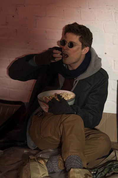 Hungry homeless man siting by brick wall and eating popcorn — Stock Photo