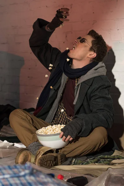 Hambriento sin hogar hombre comer palomitas de maíz mientras está sentado por la pared de ladrillo - foto de stock