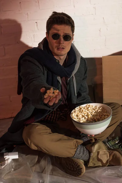 Homeless man holding popcorn in raised hand while sitting by white brick wall — Stock Photo