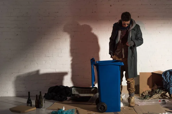 Hombre sin hogar de pie cerca de contenedor de basura rodeado de basura - foto de stock