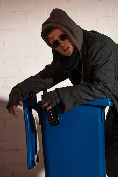 Homeless man with alcohol bottle leaning on trash container — Stock Photo