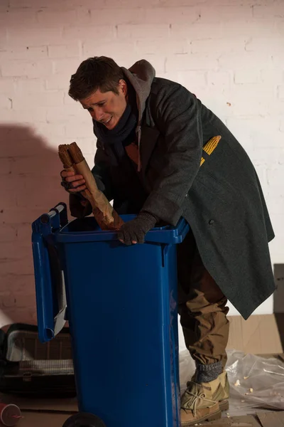 Hungry homeless man getting bread baguette from trash container — Stock Photo