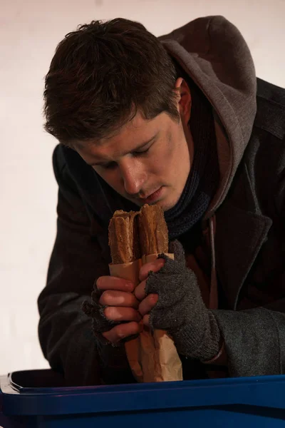 Hungry homeless man holding bread baguette while leaning on trash container — Stock Photo