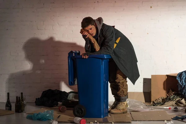 Hombre sin hogar sosteniendo pan baguette cerca de la cara mientras se apoya en contenedor de basura - foto de stock
