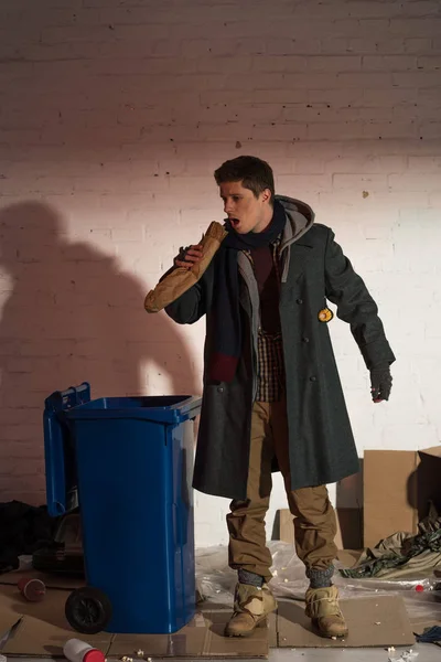 Hungry homeless man eating bread baguette while standing by trash container surrounded by rubbish — Stock Photo