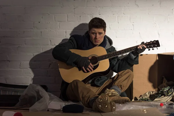 Triste hombre sin hogar tocando la guitarra acústica cerca de la pared de ladrillo blanco - foto de stock