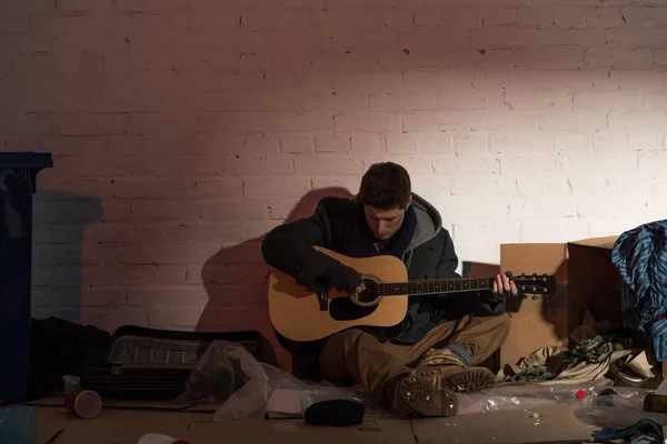 Hombre sin hogar tocando la guitarra mientras está sentado rodeado de basura - foto de stock