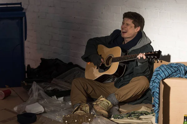 Hombre sin hogar tocando la guitarra y cantando mientras está sentado en el basurero - foto de stock