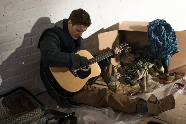 Hombre sin hogar sosteniendo la guitarra mientras está sentado cerca de la pared de ladrillo rodeado de basura - foto de stock