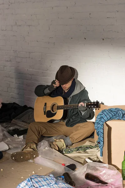Бездомный в шляпе и перчатках без пальцев держит гитару, сидя в окружении мусора — стоковое фото