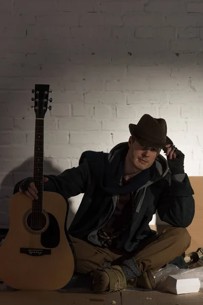 Hombre sin hogar en sombrero sentado en cartón por la pared de ladrillo blanco y la celebración de la guitarra — Stock Photo