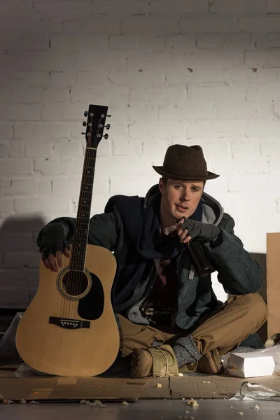 Бездомний чоловік тримає гітару, сидячи на картоні в оточенні сміття — стокове фото
