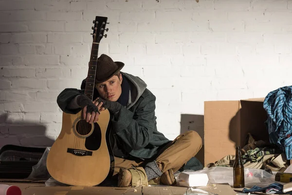 Deprimido sin hogar hombre apoyándose en la guitarra mientras se sienta en la calle basurero - foto de stock