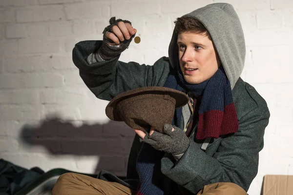 Homeless man in hood throwing coin into hat — Stock Photo