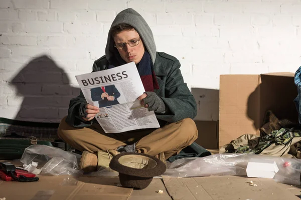 Mendigo sin hogar leyendo periódico de negocios mientras está sentado junto a la pared de ladrillo - foto de stock