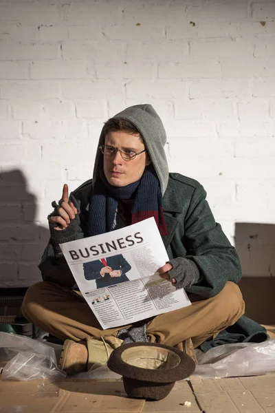Hombre sin hogar leyendo periódico de negocios y mostrando signo de idea - foto de stock