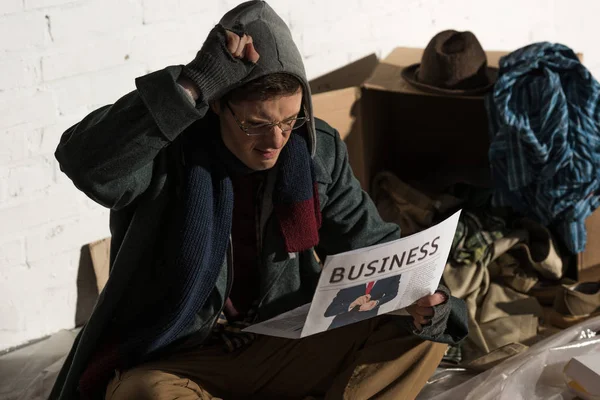 Постраждалий безпритульний чоловік читає бізнес-газету, сидячи в оточенні сміття — стокове фото