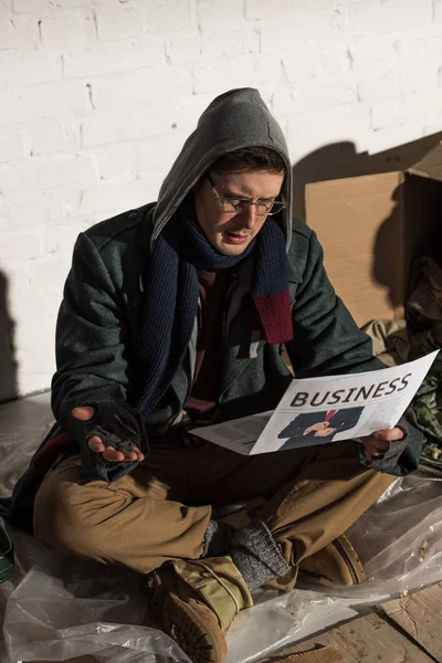 Sorprendido hombre sin hogar leyendo periódico de negocios mientras estaba sentado en el basurero - foto de stock