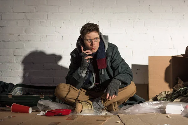 Hombre sin hogar usando teléfono vintage mientras está sentado en el vertedero de basura - foto de stock