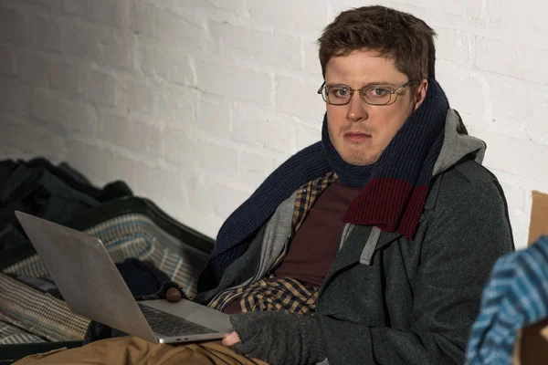 Homeless man in glasses using laptop while sitting by white brick wall — Stock Photo