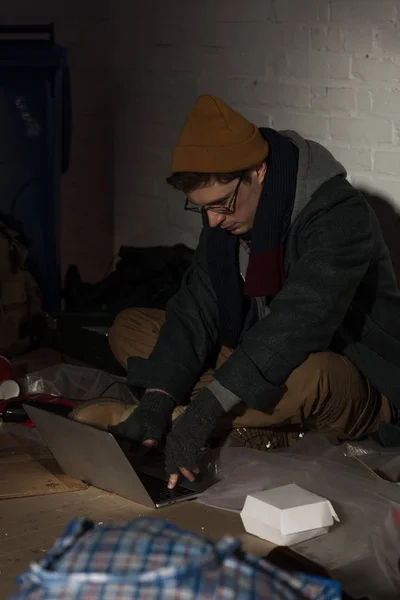 Бездомний чоловік використовує ноутбук, сидячи біля білої цегляної стіни в оточенні сміття — стокове фото