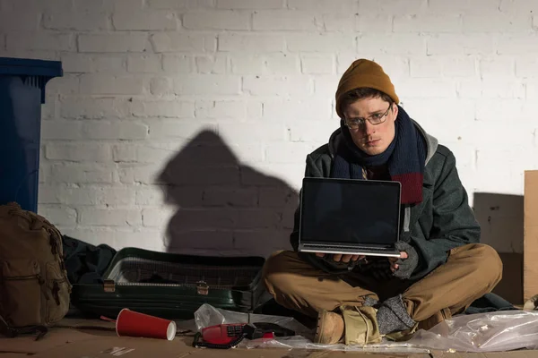 Serious homeless man using laptop while sitting by brick wall on rubbish dump — Stock Photo