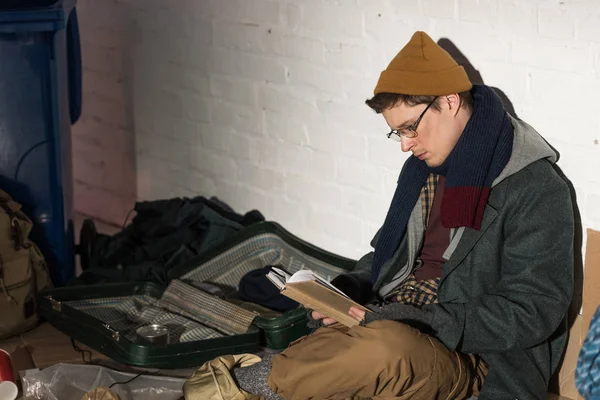 Obdachloser mit Brille liest Buch, während er auf Müllhalde sitzt — Stockfoto