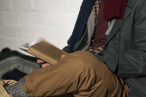 Cropped view of homeless man reading book while sitting by white brick wall — Stock Photo