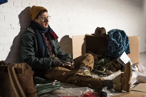 Homeless man sitting by brick wall surrounded by rubbish — Stock Photo