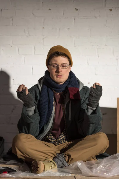 Homeless man sitting in lotus pose with closed eyes near white brick wall — Stock Photo