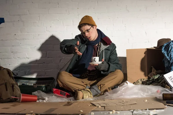 Hombre sin hogar sosteniendo taza de café y el uso de teléfono vintage mientras se sienta en el vertedero de basura - foto de stock