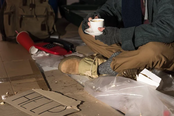 Обрезанный вид бездомного, держащего чашку кофе, сидящего в окружении мусора — стоковое фото