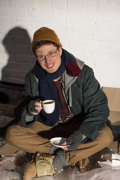 Un sans-abri souriant buvant du café assis près d'un mur de briques blanches entouré de déchets — Photo de stock