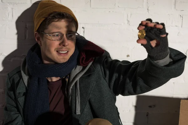 Sonriente hombre sin hogar en gafas mirando la moneda en la mano - foto de stock