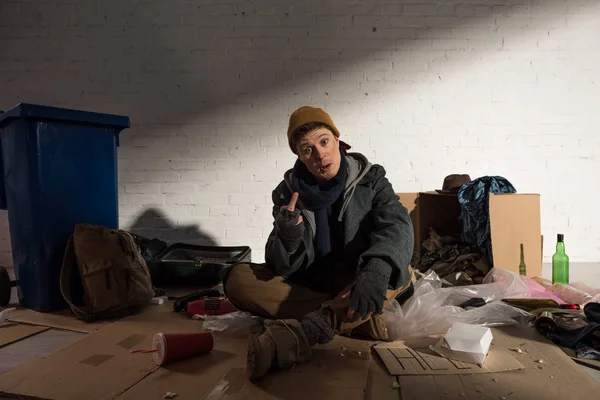 Hombre sin hogar mostrando el dedo medio mientras está sentado en el vertedero de basura - foto de stock