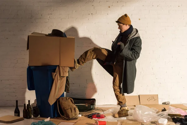 Разгневанный бездомный пинает мусорный контейнер картонной коробкой — стоковое фото