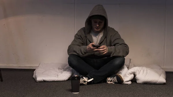 Человек в толстовке с помощью смартфона, сидя на матрасе расположены на полу — стоковое фото