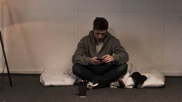 Homme en utilisant smartphone tout en étant assis sur un matelas disposé sur le sol dans la chambre noire — Photo de stock