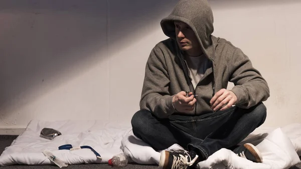 Addictive man in hoodie holding smoking pipe while sitting on mattress in dark room — Stock Photo