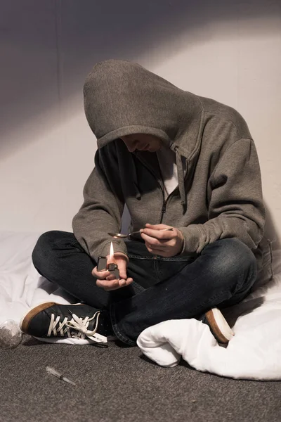 Addict man seating on bedding and heating spoon with heroin on lighter — Stock Photo