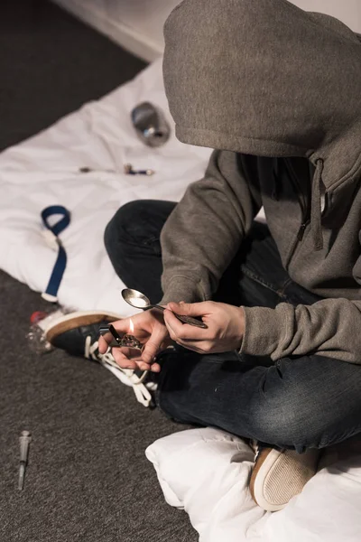 Junkie man in hood heating spoon with heroin on lighter — Stock Photo