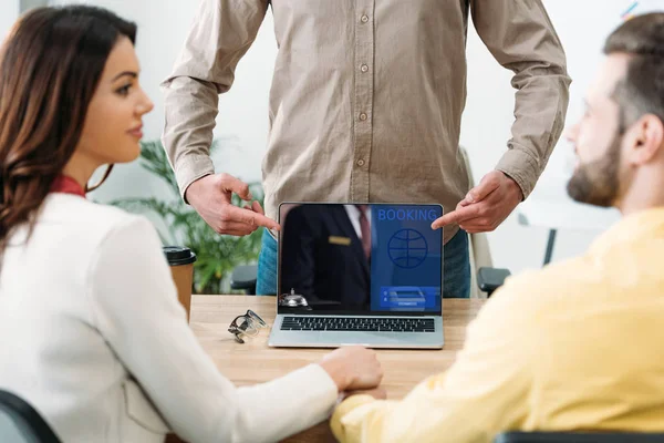 Selektiver Fokus des Beraters zeigt mit dem Finger auf Laptop mit Buchungswebseite auf dem Bildschirm in der Nähe des Ehepaares im Büro — Stockfoto