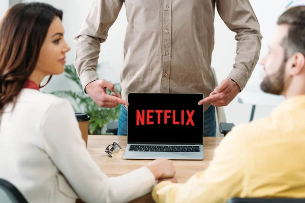 Selective focus of advisor pointing with fingers at laptop with netflix website on screen to couple in office — Stock Photo