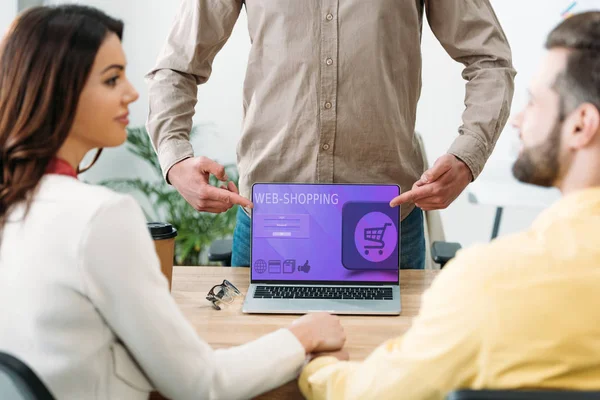 Selective focus of advisor pointing with fingers at laptop with web shopping website on screen to couple in office — Stock Photo