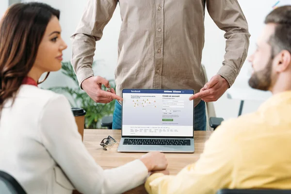 Selective focus of advisor pointing with fingers at laptop with facebook website on screen to couple in office — Stock Photo