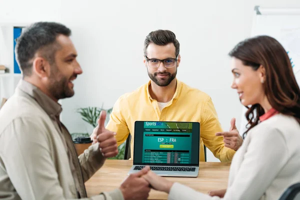 Selective focus of advisor pointing with fingers at laptop with sportsbet online website on screen to couple in office — Stock Photo