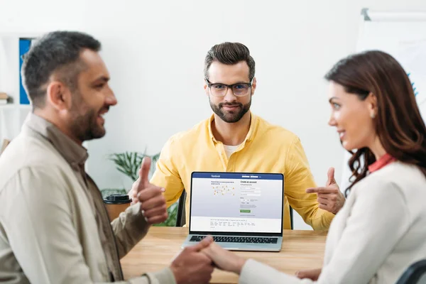 Selective focus of advisor pointing with finger at laptop facebook website on screen while investor holding woman hand and thumbing up in office — Stock Photo