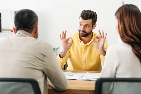 Messa a fuoco selettiva del consulente seduto a tavola e mostrando segno okey per l'uomo e la donna in carica — Foto stock