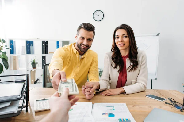 Enfoque selectivo de la pareja de inversores sentados en la mesa y tomando billetes de dólar de asesor en el cargo - foto de stock