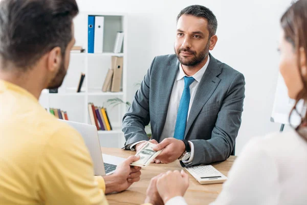 Selective focus of advisor sitting at table and giving dollar banknote to couple investors in office — Stock Photo